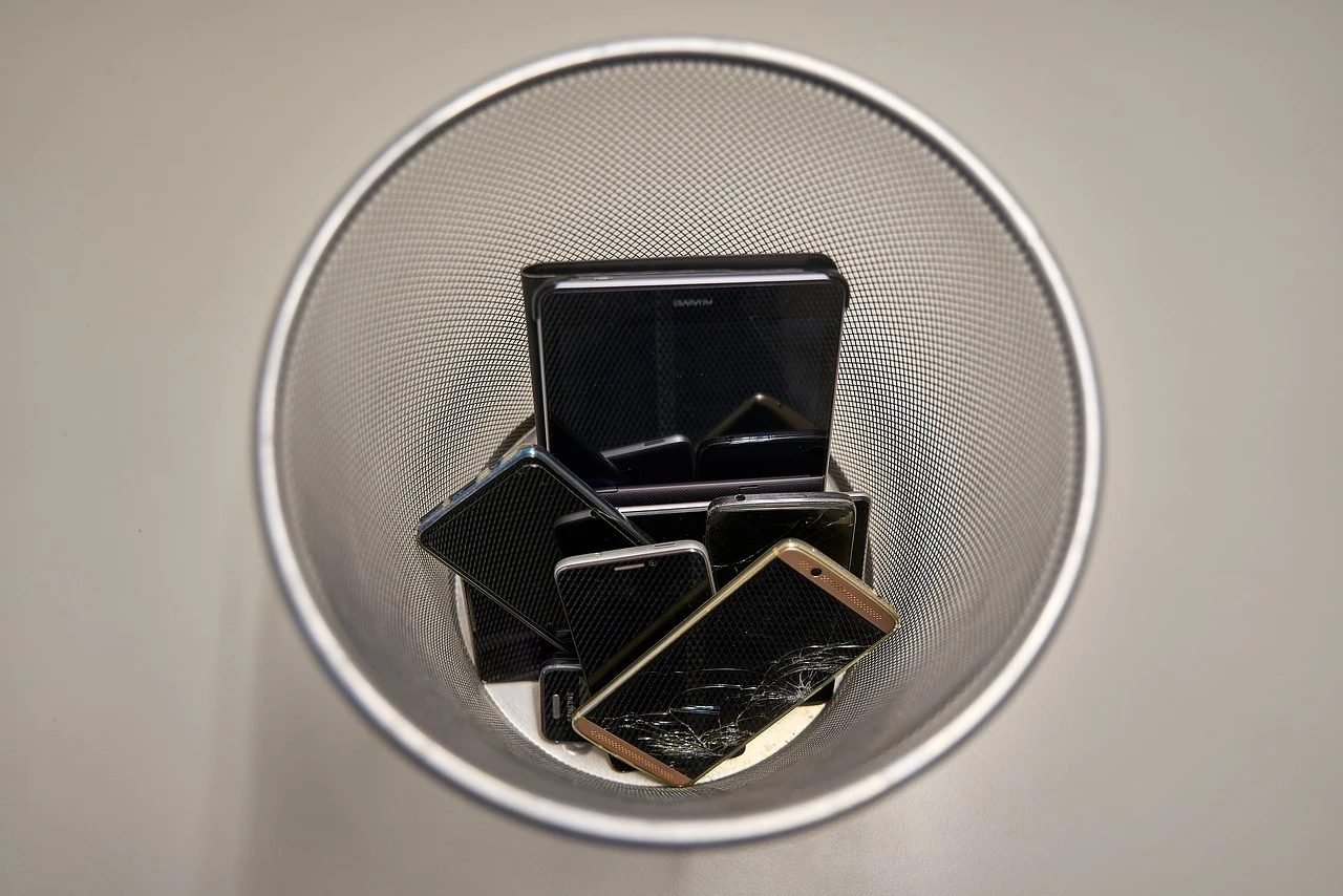 Kosz na śmieci z kilkoma starymi smartfonami wewnątrz, widziany z góry. Telefony są ułożone w nieładzie, co sugeruje ich nieużyteczność lub przeznaczenie do recyklingu.