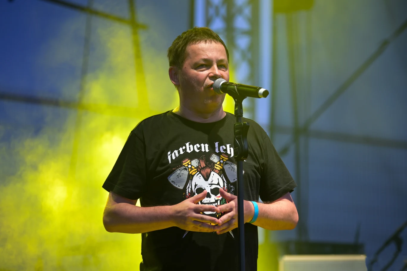 Wojtek Wojda stoi na czele Farben Lehre Mężczyzna w koszulce z grafiką przemawia do mikrofonu na tle sceny oświetlonej żółtymi reflektorami.