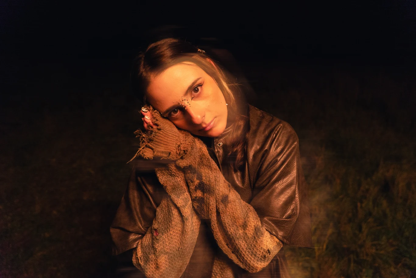 Osoba z długimi, ciemnymi włosami spogląda intensywnie w obiektyw, opierając głowę na rękach ubranych w grube, wełniane rękawice. Scena oświetlona ciepłym, miękkim światłem na tle ciemnego otoczenia.