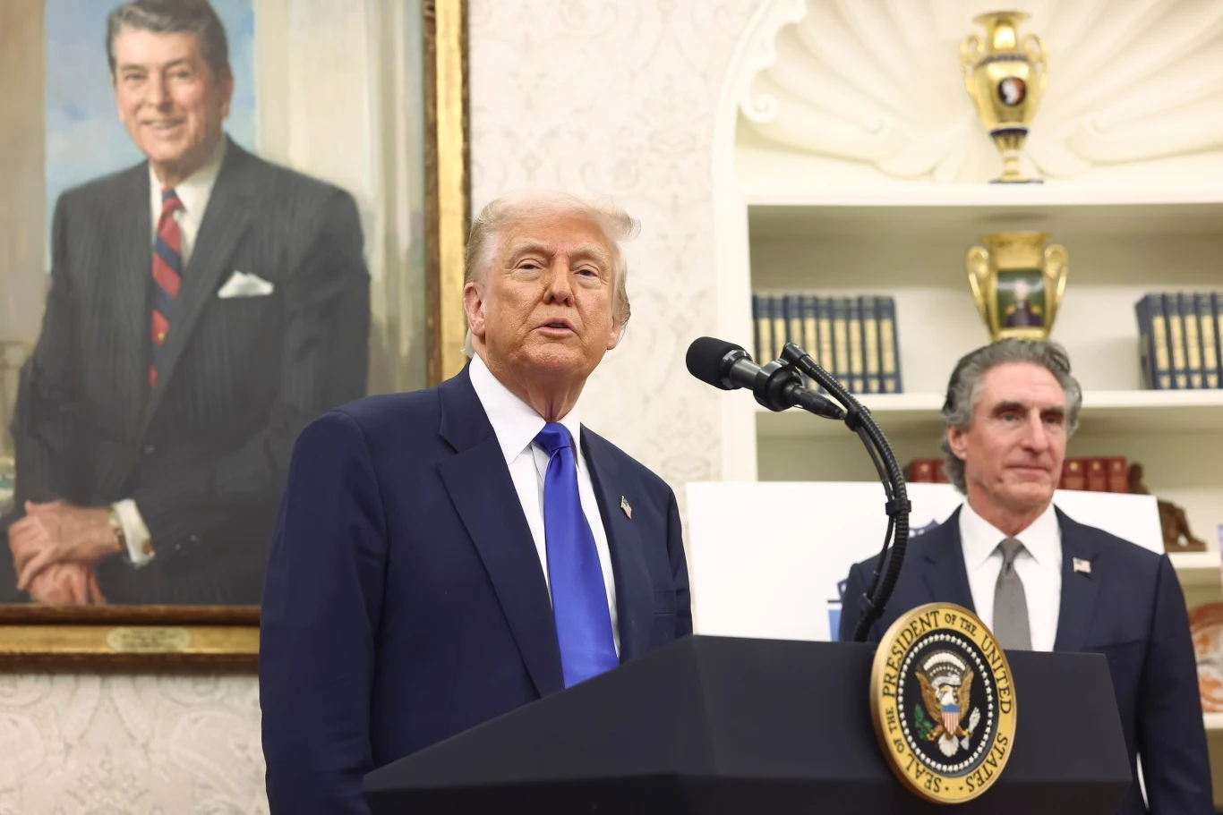 Prezydent USA Donald Trump Dwóch mężczyzn stoi przed mikrofonem w eleganckich garniturach. Na pierwszym planie znajduje się mównica z godłem prezydenckim USA. W tle duży portret mężczyzny w garniturze oraz półki z książkami.