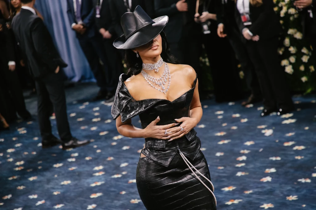 Kapelusz Kim Kardashian dodawał jej tajemniczości Kobieta w eleganckiej czarnej sukni i kapeluszu stoi na dywanie w tłocznym miejscu, otoczona przez innych ludzi w formalnych strojach. Ma na sobie wyrazistą biżuterię, która przyciąga uwagę.