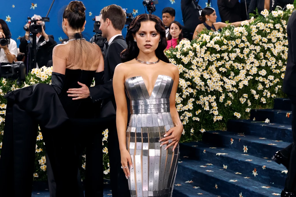 Jenna Ortega zaskoczyła materiałem swojej kreacji Młoda kobieta w srebrnej, błyszczącej sukni stoi na niebieskim dywanie, otoczona kwiatami i innymi uczestnikami eleganckiego wydarzenia.