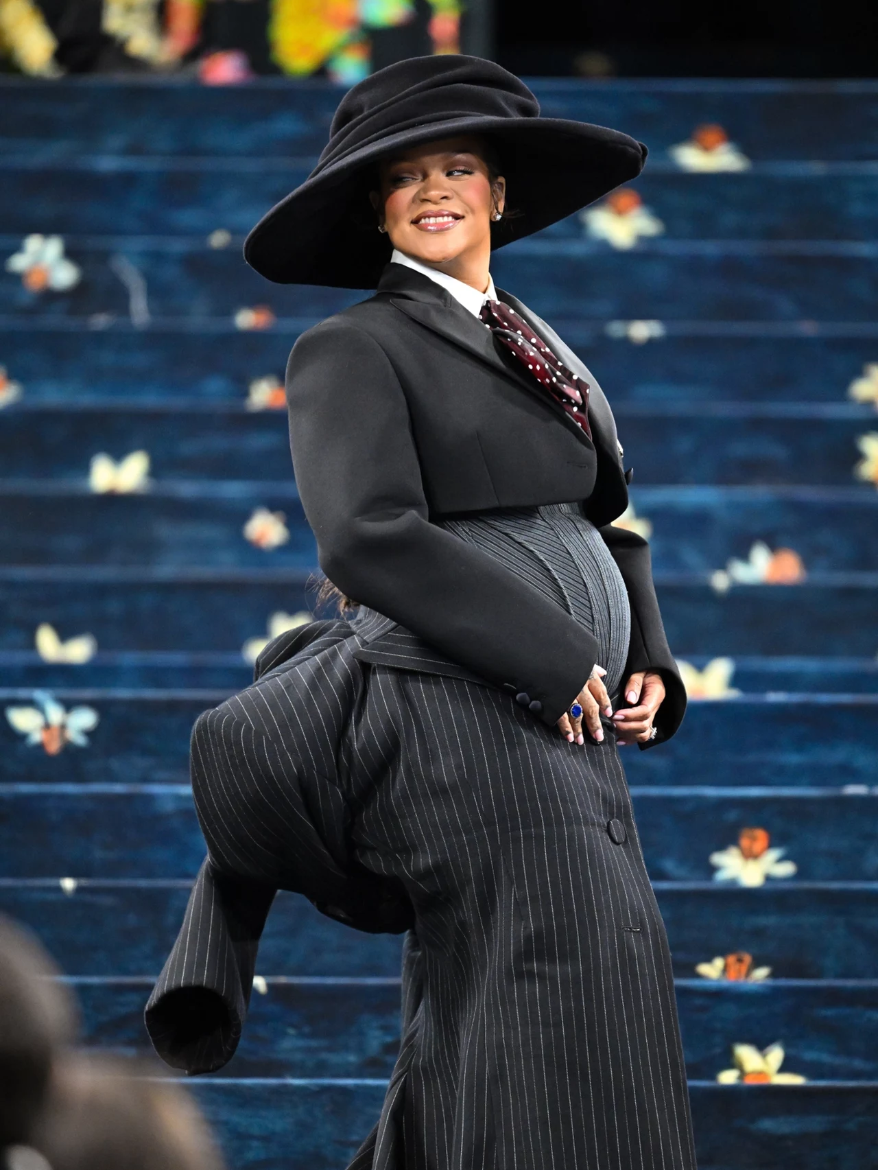 Rihanna swoim pojawieniem się na Met Gali potwierdziła kolejną ciążę Osoba ubrana w elegancki, czarny garnitur w prążki oraz dużą kapelusz czarnym tle z błękitnymi schodami ozdobionymi motywem kwiatowym. Osoba delikatnie obejmuje dłońmi zaokrąglony brzuch, co sugeruje ciążę.
