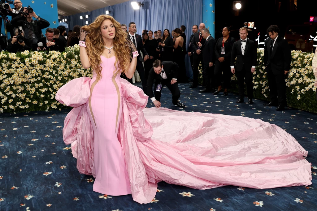 Shakira zaskoczyła samą swoją obecnością i rozmiarem trenu swojej sukni Osoba w efektownej różowej sukni z długim trenem na czerwonym dywanie, otoczona przez fotografów i gości wydarzenia. Tło wypełniają elegancko ubrani ludzie i dekoracje z kwiatów.