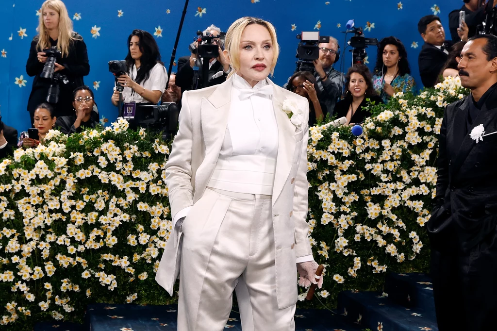 Madonna zdecydowała się na męski look i zamiast torebki w ręku trzymała cygaro Kobieta ubrana w elegancki biały garnitur stoi na tle kwiatowej dekoracji, w otoczeniu licznych fotoreporterów i kamerzystów. W tle widać niebieską ścianę ozdobioną małymi gwiazdkami, która dodaje scenie wyjątkowy charakter.