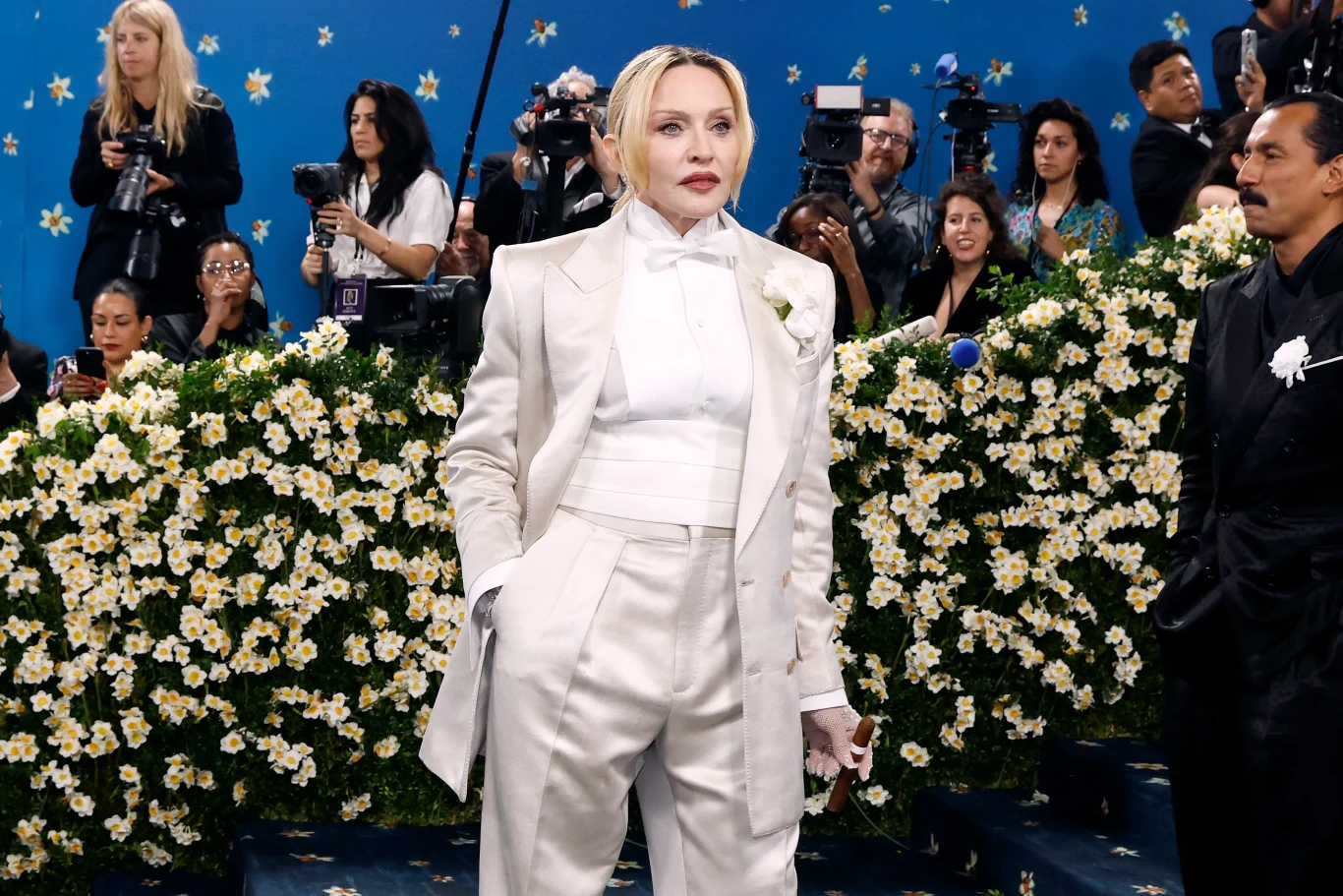Madonna zdecydowała się na męski look i zamiast torebki w ręku trzymała cygaro Kobieta ubrana w elegancki biały garnitur stoi na tle kwiatowej dekoracji, w otoczeniu licznych fotoreporterów i kamerzystów. W tle widać niebieską ścianę ozdobioną małymi gwiazdkami, która dodaje scenie wyjątkowy charakter.