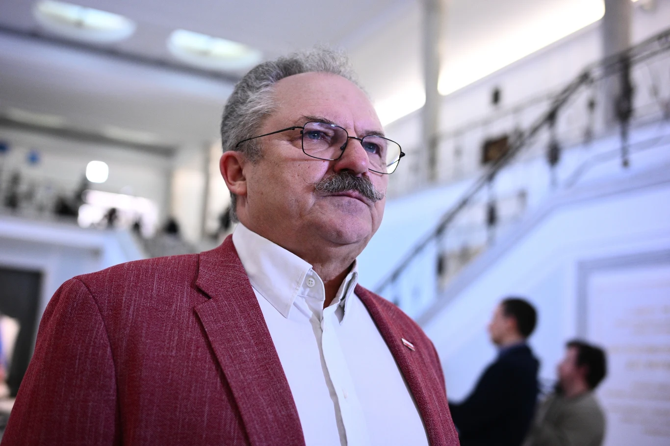 Kandydat na prezydenta RP Marek Jakubiak Mężczyzna w czerwonej marynarce stoi wewnątrz budynku o białych ścianach i eleganckich schodach. W tle widoczne są inne osoby w nieostrości, a wnętrze cechuje się nowoczesnym i stylowym wystrojem.