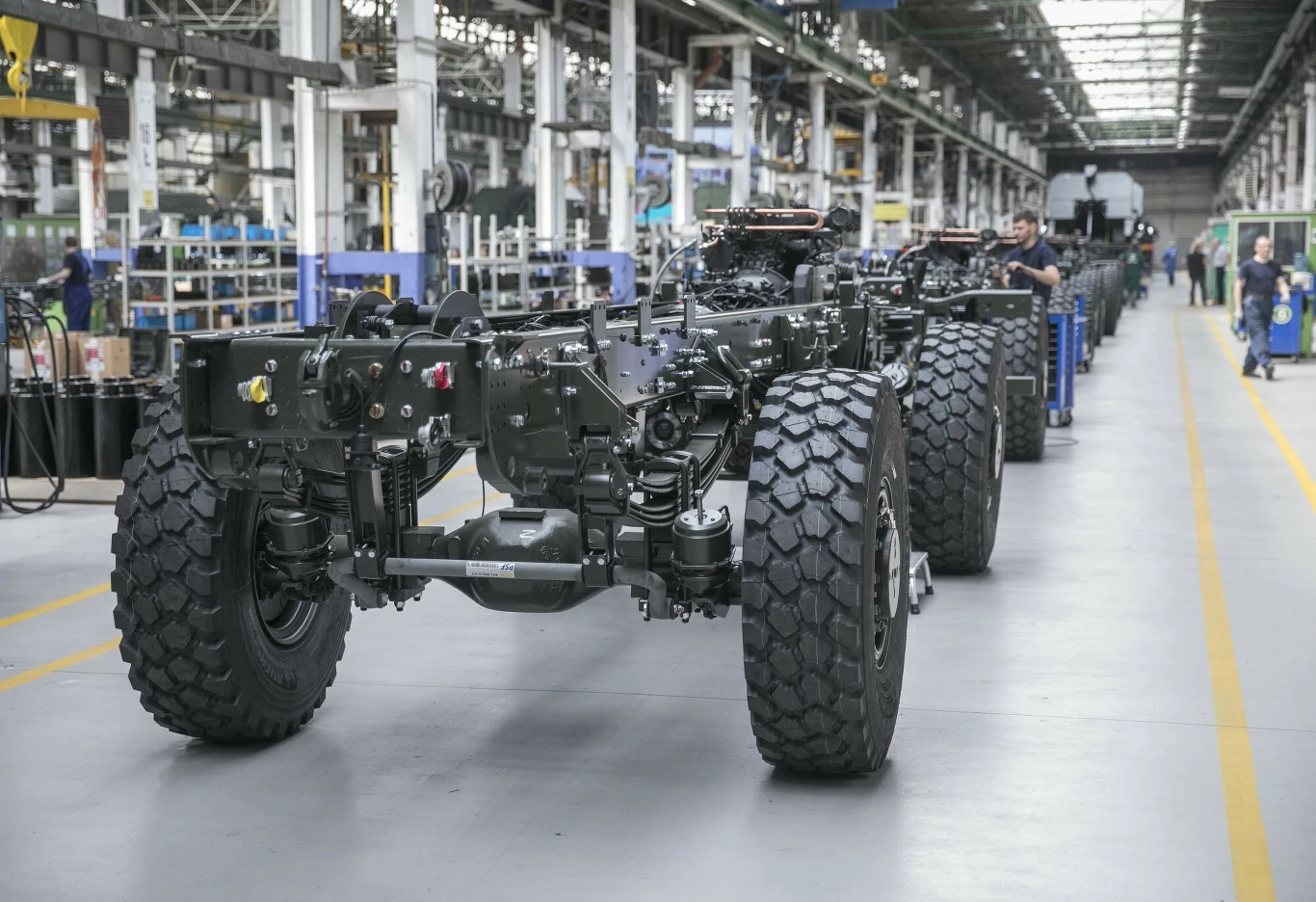 Linia produkcyjna w fabryce pojazdów, z widocznymi podwoziami na różnych etapach montażu. Przestronna hala z jasnym oświetleniem, widać skupionych pracowników w tle, koordynujących proces produkcji.