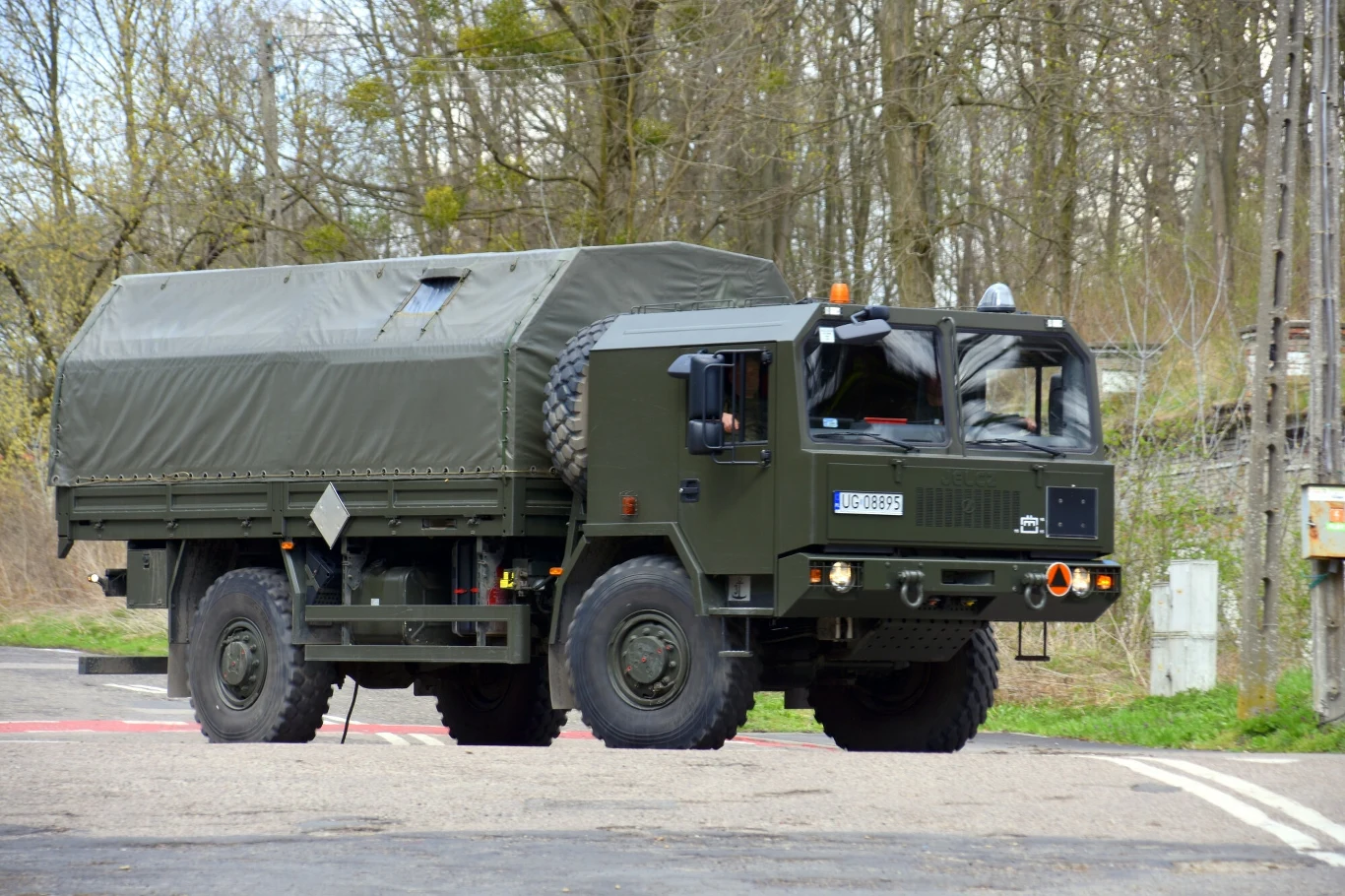 Jelcz na dobre wraca do gry. Wkrótce kolejne zamówienia dla wojska. Wojskowa ciężarówka z plandeką na pace porusza się po asfaltowej drodze, otoczona drzewami i zielenią w tle.
