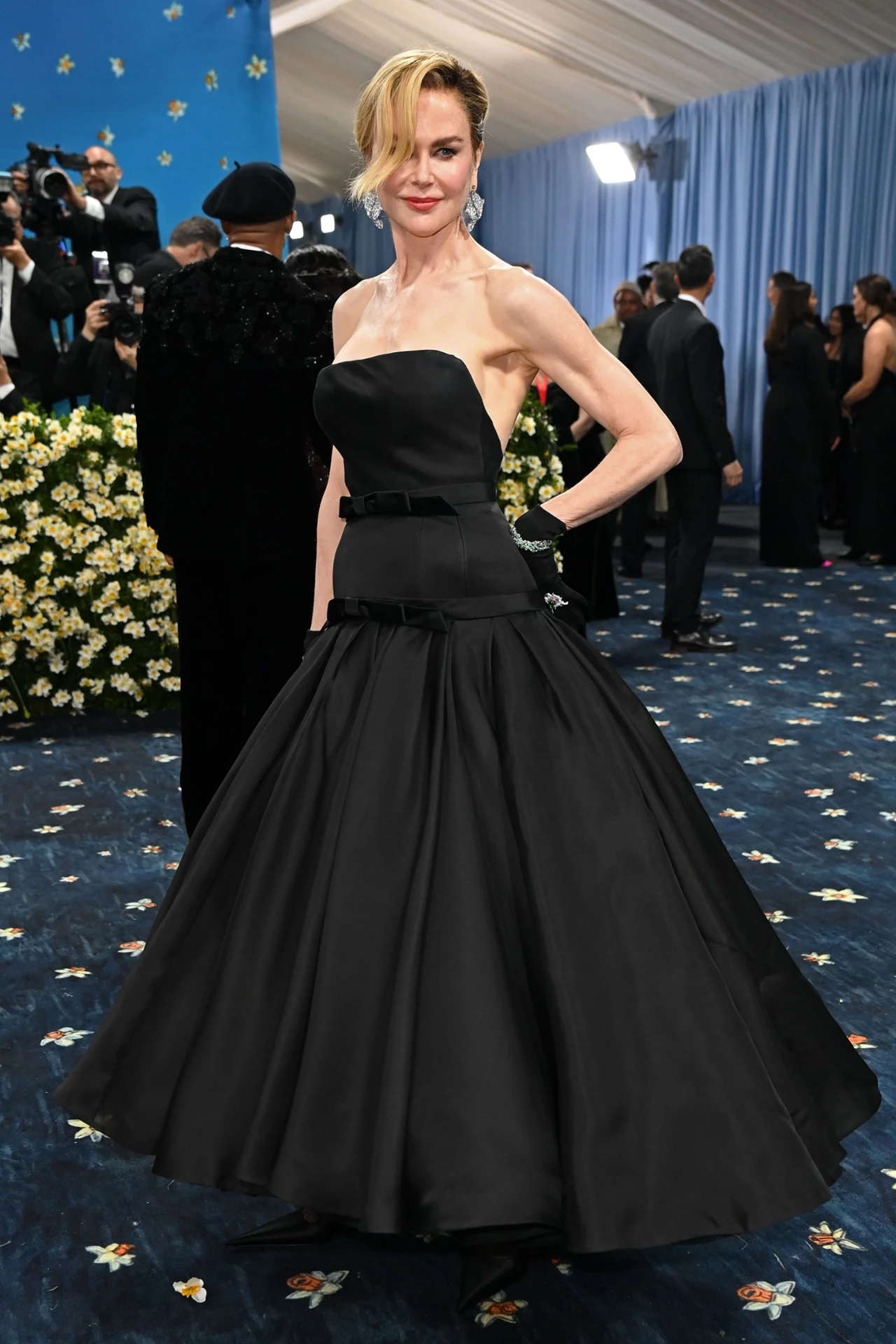 Idealnym dopełnieniem fryzury była czarna suknia od Balenciagi Kobieta ubrana w elegancką, czarną suknię wieczorową na wydarzeniu o formalnym charakterze. Tło tworzy niebieska zasłona oraz grupa osób w strojach wieczorowych.