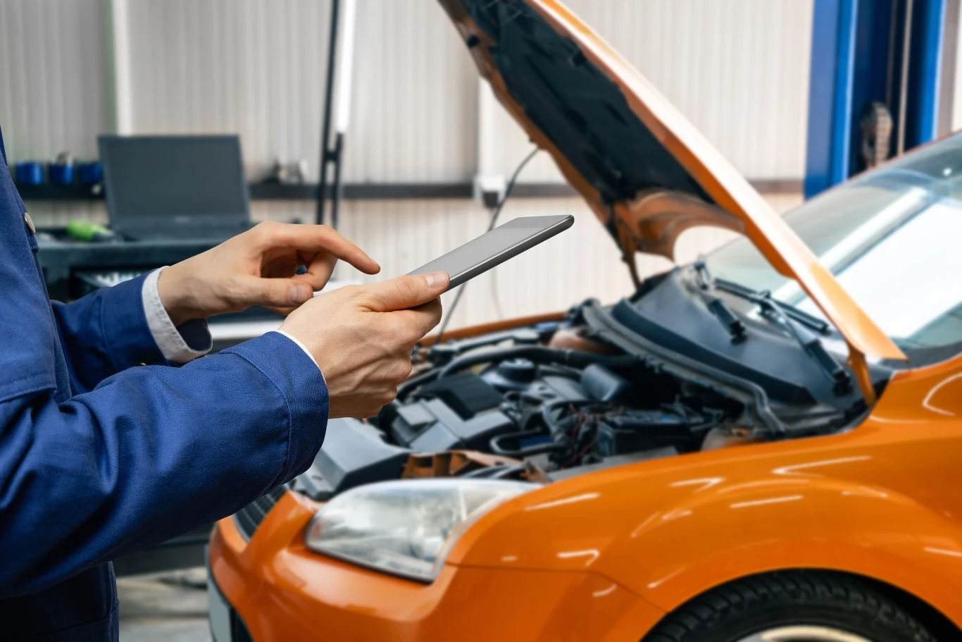 mechanik w niebieskim uniformie używający tabletu do diagnostyki pomarańczowego samochodu z otwartą maską w warsztacie samochodowym