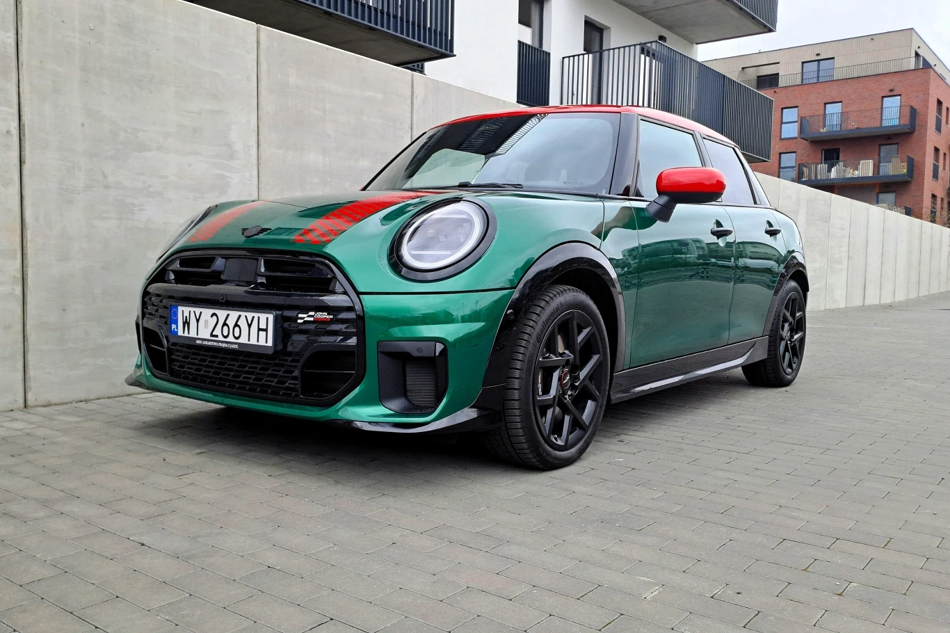 Mini Cooper był najlepiej sprzedającym się modelem brytyjskiego producenta w Polsce w 2025 roku. Zielony samochód marki Mini zaparkowany na chodniku w otoczeniu nowoczesnych budynków. Sportowy charakter pojazdu podkreślają czerwone akcenty na masce i lusterkach. Kod rejestracyjny widoczny z przodu pojazdu.