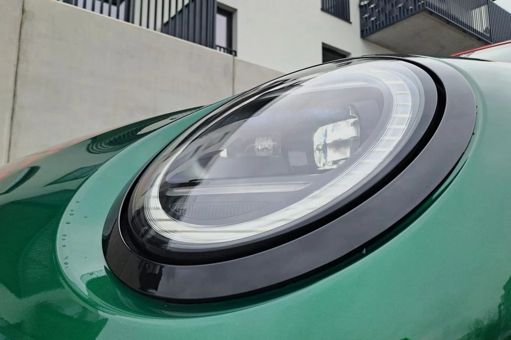 Stylistycznie spalinowy i elektryczny Cooper zbytnio się od siebie nie różnią. Znajdziemy tutaj np. takie same okrągłe światła (nawiązujące do pierwszego Mini), ale podzielone. Zbliżenie na okrągły reflektor samochodowy w nowoczesnym, zielonym samochodzie, z widocznymi detalami konstrukcyjnymi i odbiciem otoczenia w powierzchni lampy. W tle fragmenty nowoczesnego budynku mieszkalnego.