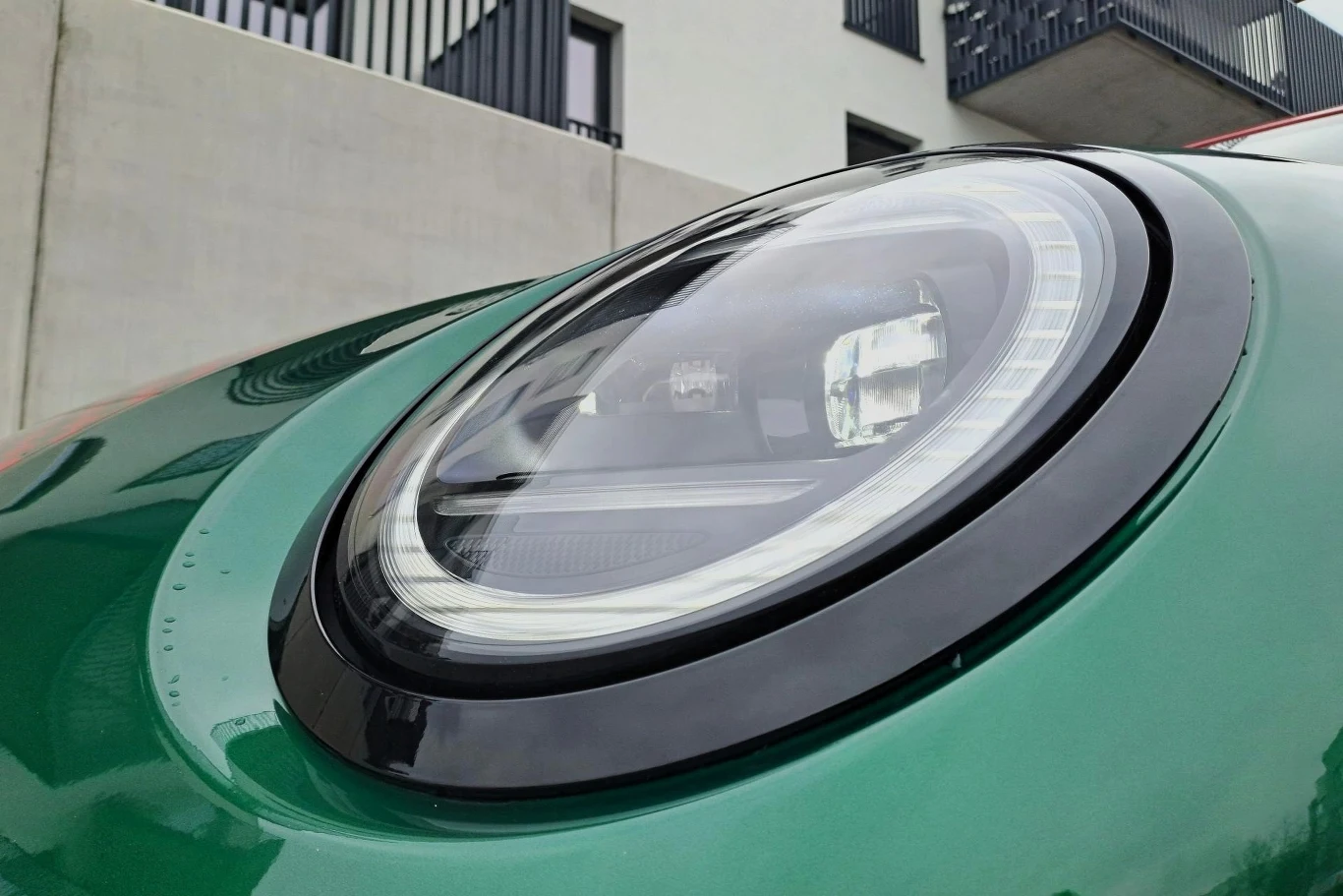 Zbliżenie na okrągły reflektor samochodowy w nowoczesnym, zielonym samochodzie, z widocznymi detalami konstrukcyjnymi i odbiciem otoczenia w powierzchni lampy. W tle fragmenty nowoczesnego budynku mieszkalnego.