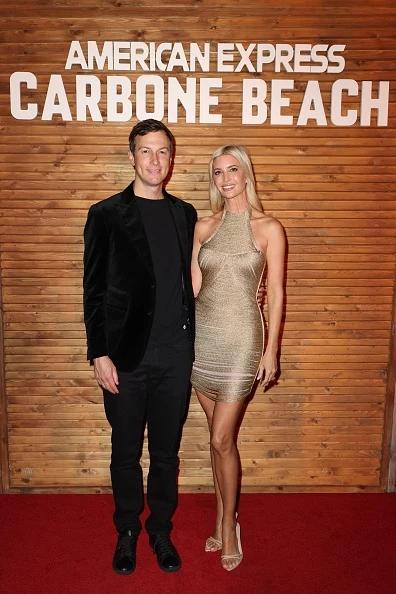 Ivanka Trump z mężem Dwie elegancko ubrane osoby stoją na czerwonym dywanie. Kobieta ubrana jest w połyskującą, srebrzystą sukienkę, a mężczyzna w czarną marynarkę i spodnie. W tle znajduje się drewniana ściana z napisem 'American Express Carbone Beach'.