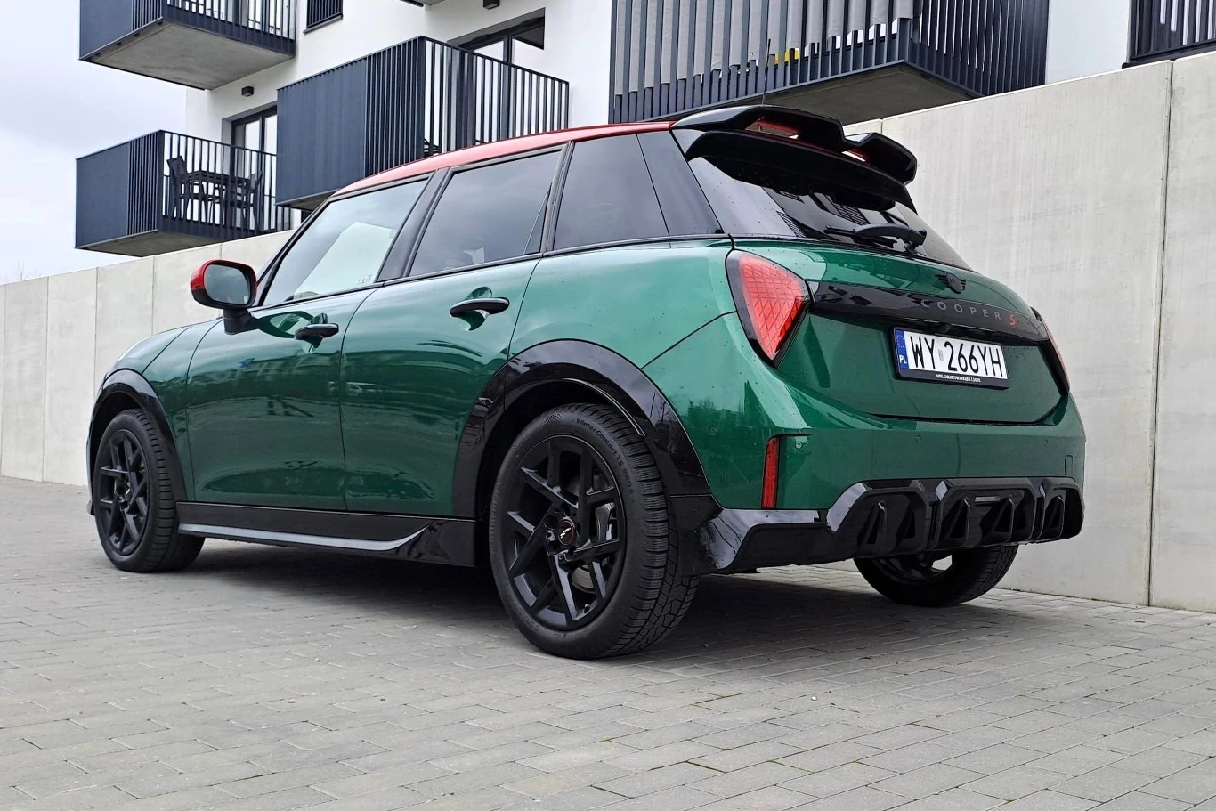 Zielony samochód zaparkowany przed nowoczesnym budynkiem z balkonami. Auto ma czerwony dach i czarne felgi, co nadaje mu sportowy wygląd. Po lewej stronie znajduje się jasna ściana, która kontrastuje z zielenią pojazdu.