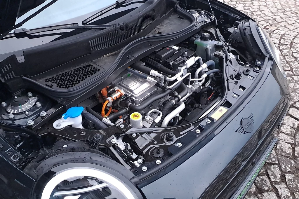 Mini Cooper w wersji elektrycznej jest dostępny w trzech wersjach napędu i dwóch wariantach akumulatora - w standardzie 36,6 kWh, a w dwóch wyższych 49,2 kWh. Otwarta maska samochodu elektrycznego z widocznym silnikiem i elementami technicznymi, w tym przewody i zbiorniki płynów.