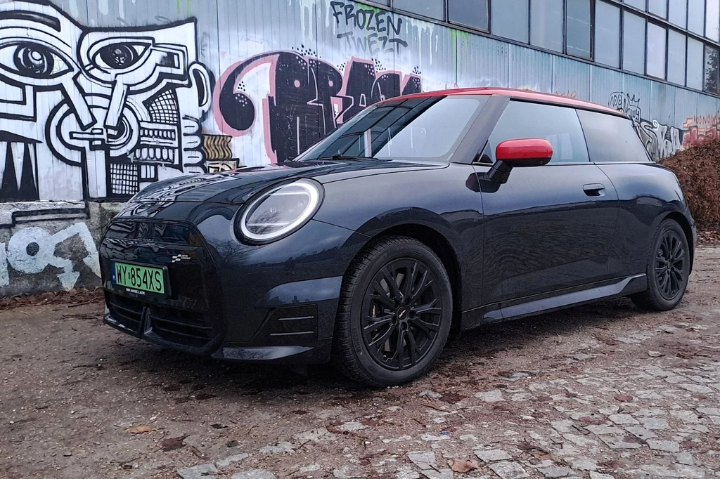 Ceny elektrycznego Coopera startują od 146 500 zł. Wszystkie warianty objęte są dopłatami z programu "NaszEauto". Czarny samochód Mini Cooper zaparkowany na brukowanej ulicy przed ścianą pokrytą różnorodnymi graffiti. W tle widoczne są duże okna budynku.