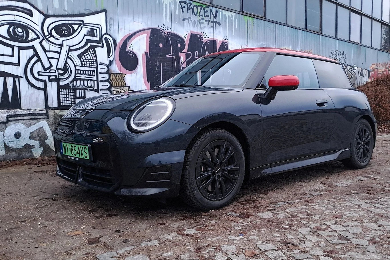 Czarny samochód Mini Cooper zaparkowany na brukowanej ulicy przed ścianą pokrytą różnorodnymi graffiti. W tle widoczne są duże okna budynku.