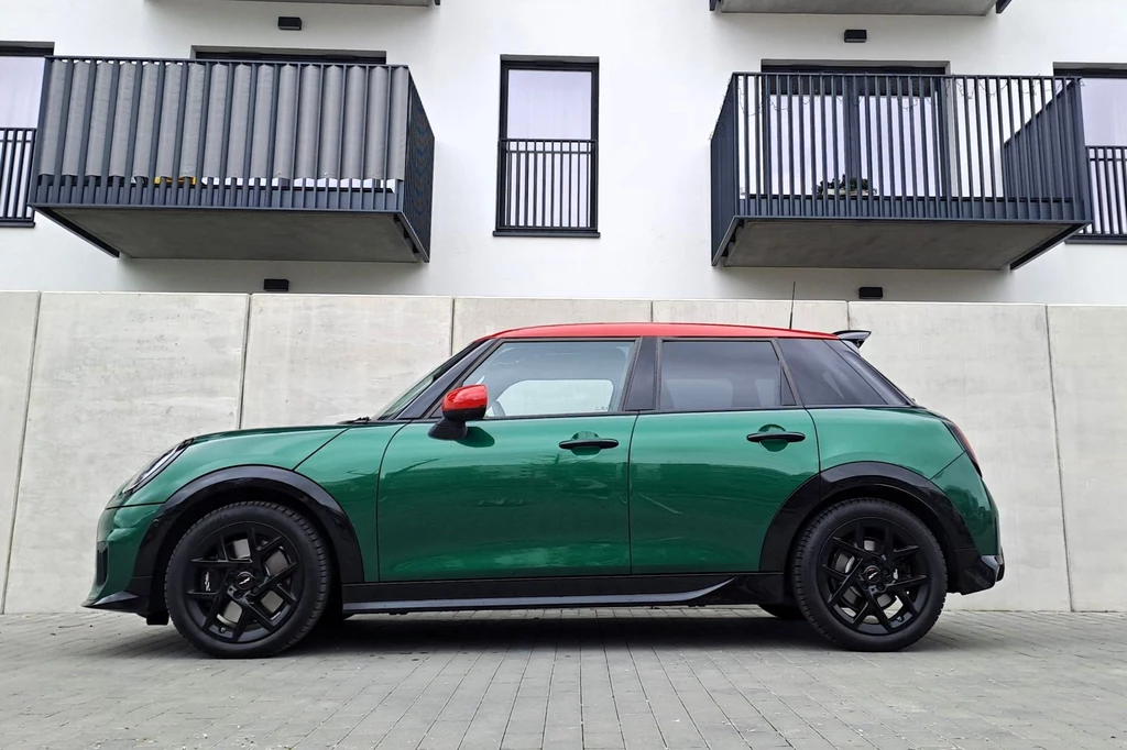Spalinowy Mini Cooper w podstawowej specyfikuje kosztuje 111 900 zł (wersja trzydrzwiowa) lub 115 900 zł (wersja pięciodrzwiowa). Zielony samochód o nowoczesnym designie zaparkowany przed budynkiem z szarymi balkonami. Samochód ma czarną felgę, czerwony dach i lusterka boczne. Tło stanowią betonowe ściany i nowoczesny budynek mieszkalny.