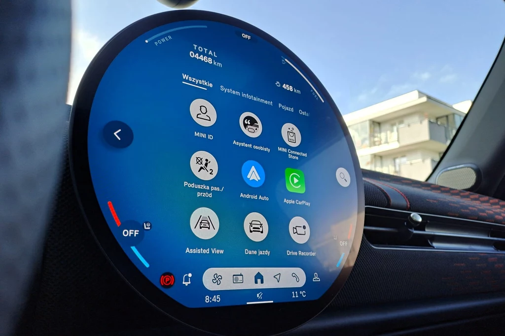 Zarówno w wariancie elektrycznym, jak i spalinowym centralne miejsce zajmuje okrągły ekran. W zależności od wariantu napędu pokazuje on stan naładowania akumulatora lub poziom paliwa w zbiorniku. Kokpit samochodu z wyświetlaczem dotykowym o okrągłym kształcie, wyświetlającym ikony związane z systemem informacyjnym i funkcjami pojazdu. W tle widać nieostro zarysowany budynek.