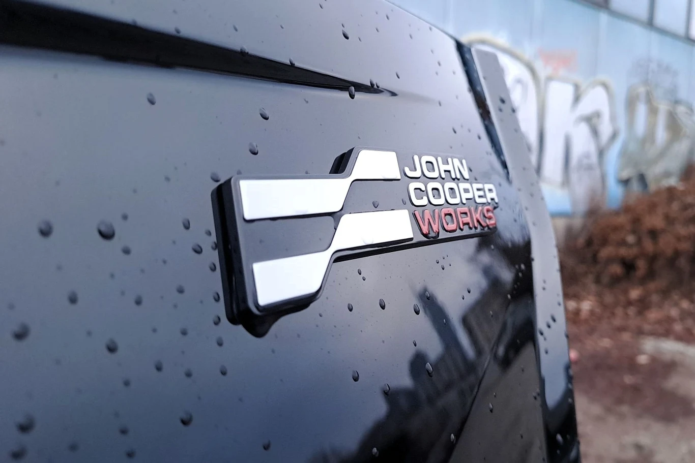 Logo John Cooper Works na czarnej karoserii samochodu pokrytej kroplami deszczu, w tle widać graffiti na ścianie budynku.