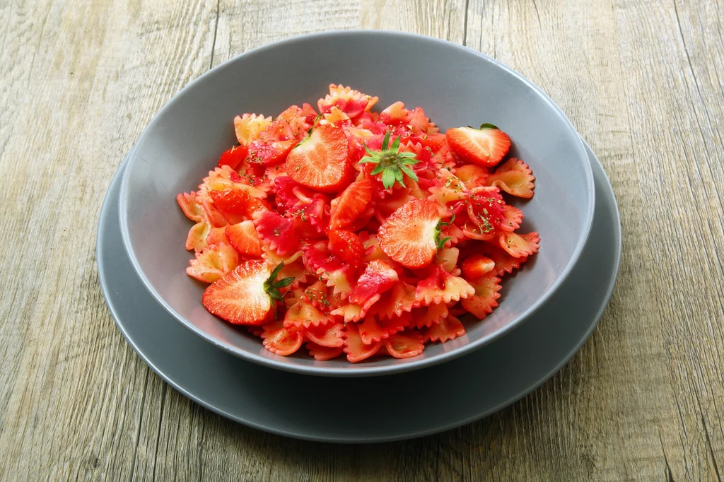 Do przygotowania tego dania można użyć każdego rodzaju makaronu Pasta farfalle w sosie truskawkowym, udekorowana świeżymi truskawkami i gałązką zielonej pietruszki, podana na szarym talerzu na drewnianym stole.