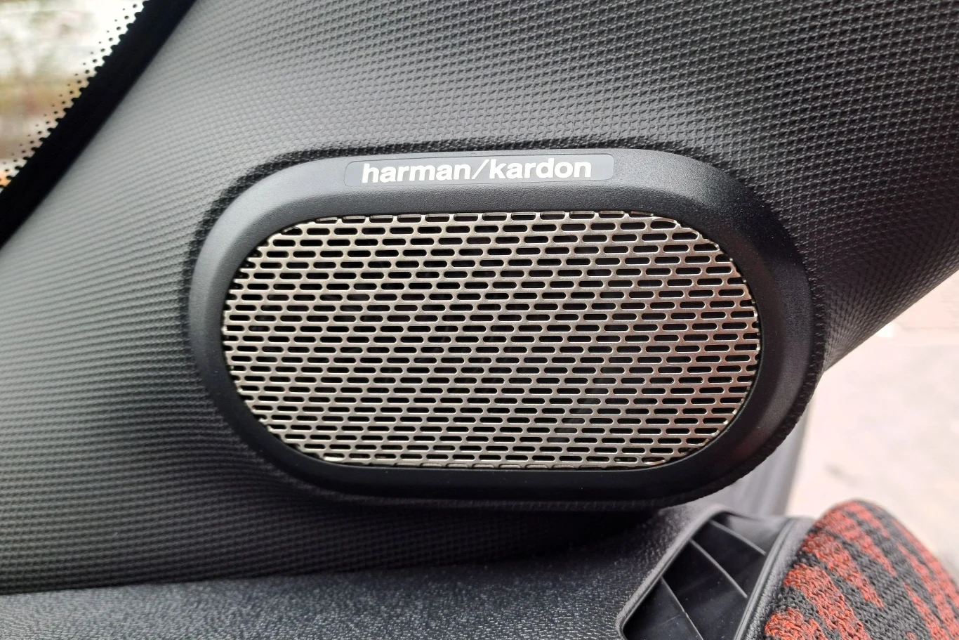 Głośnik samochodowy marki Harman Kardon zamontowany na bocznej ścianie wnętrza samochodu, wykończony w kolorze czarnym z metalową siatką ochronną.