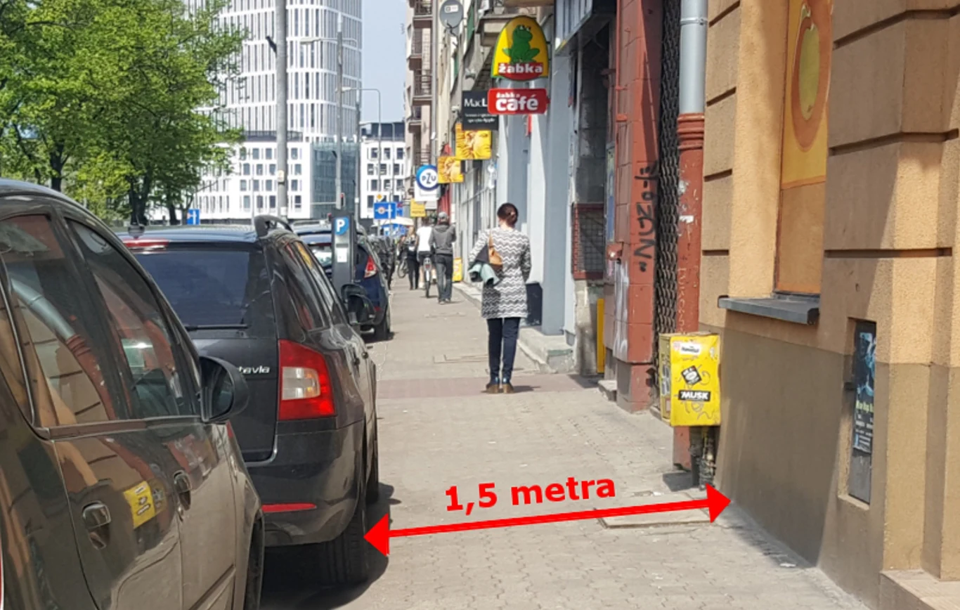 Samochód zaparkowany na chodniku, ograniczający przestrzeń do chodzenia pieszym. Ulica w tle z licznymi sklepami i reklamami oraz osoba idąca w oddali. Czerwona linia wskazuje odległość 1,5 metra między samochodem a ścianą budynku.