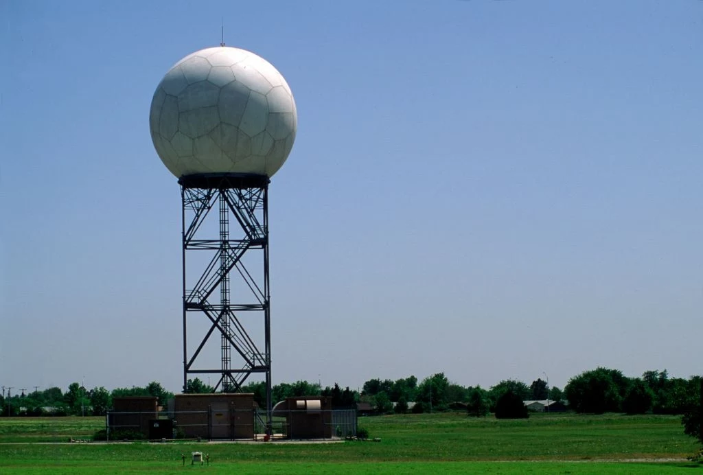 Dopplerowski radar pogodowy w Amarillo w Teksasie Stacja radarowa z dużą, kulistą kopułą osadzona na wysokiej metalowej wieży, otoczona zielonymi polami z widocznymi w oddali drzewami i budynkami.