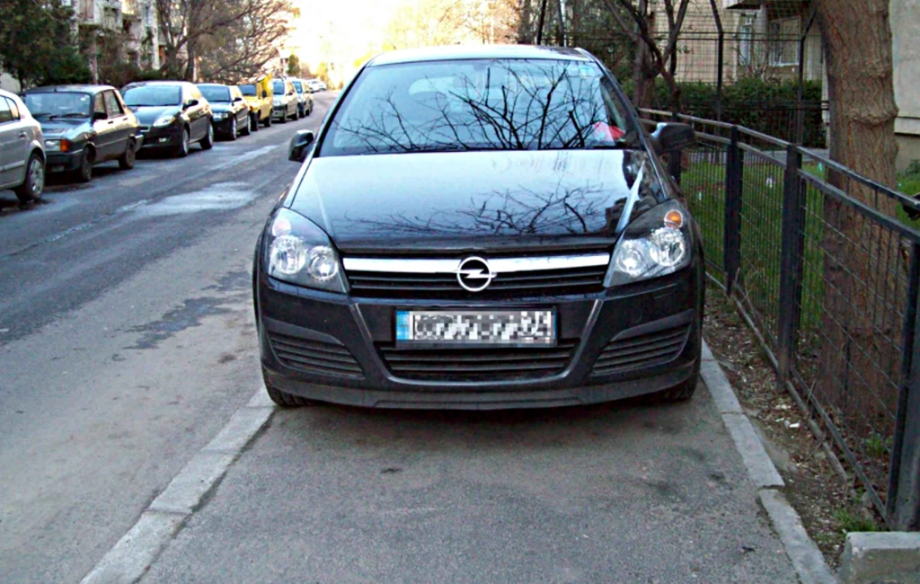 Zajmowanie całego chodnika przez samochód jest niedozwolone Samochód marki Opel zaparkowany na chodniku wzdłuż ulicy. Widoczne są inne pojazdy zaparkowane na ulicy oraz fragment budynków i drzew. Rejestracja samochodu jest nieczytelna.