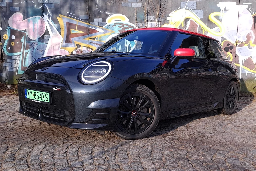 Elektryczny Mini Cooper, którego mieliśmy okazję testować, to odmiana SE. Oferuje 218 KM i 330 Nm. Czarny samochód Mini Cooper z czerwonym dachem na tle kolorowego graffiti na kamienistej ulicy.