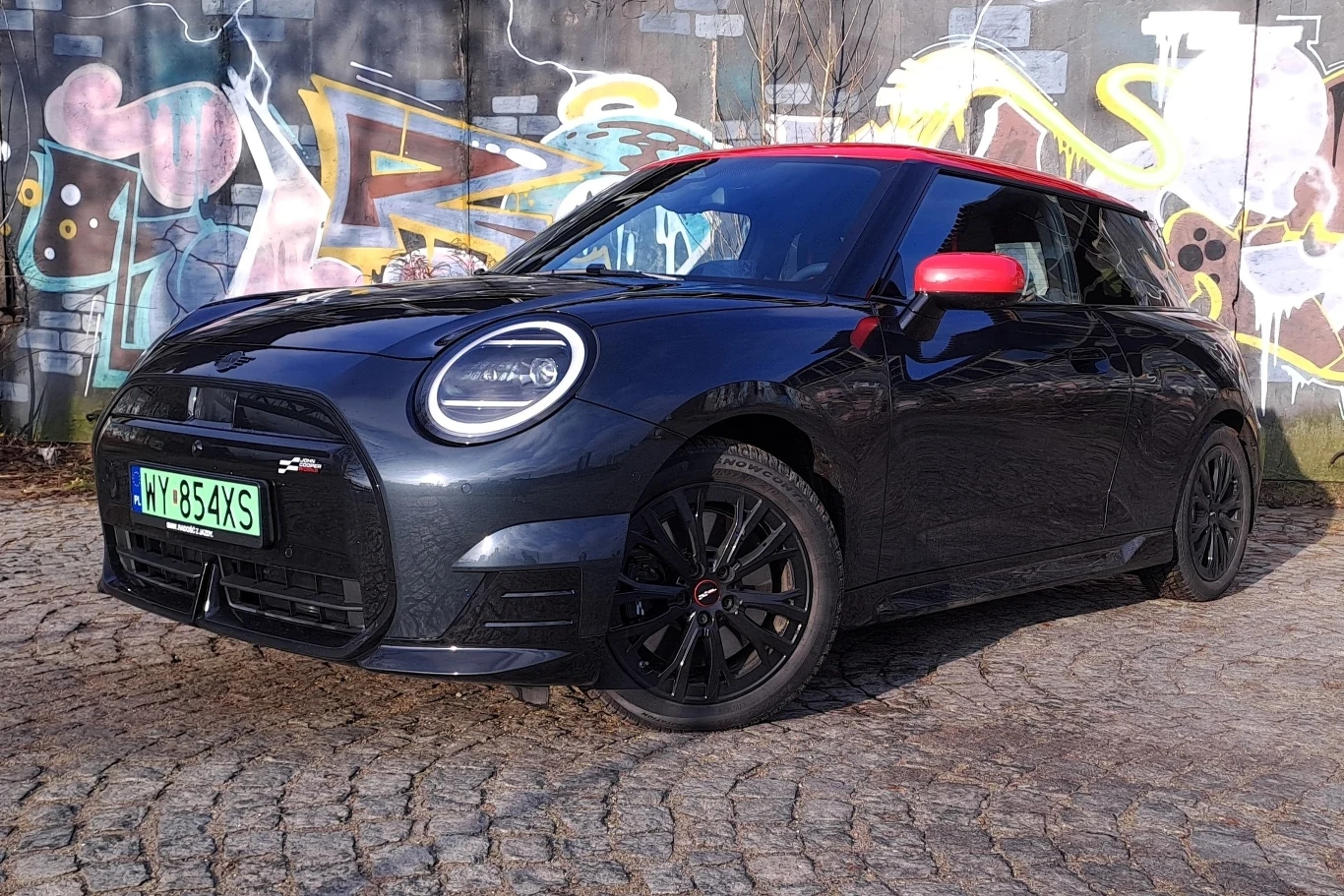 Czarny samochód Mini Cooper z czerwonym dachem na tle kolorowego graffiti na kamienistej ulicy.