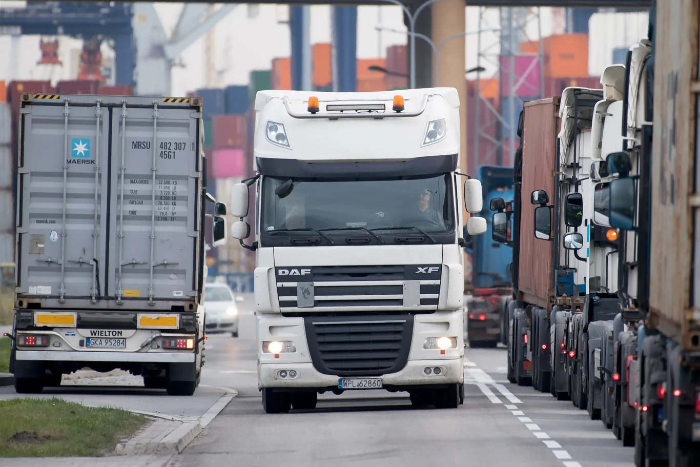 Branżę transportową czekają trudne czasy. Nowe obowiązki i opłaty Ruchliwe skrzyżowanie w porcie, ciężarówki przewożą kontenery, w tle widać dźwigi i stosy kontenerów.