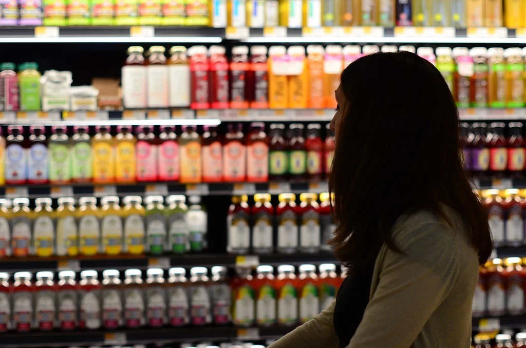 Fot. pixabay.com/ kc0uvb kobieta robiąca zakupy w supermarkecie, stojąca przed półkami pełnymi różnokolorowych butelek z napojami; nieprzejrzyste zdjęcie z widocznymi konturami półek i produktów w tle