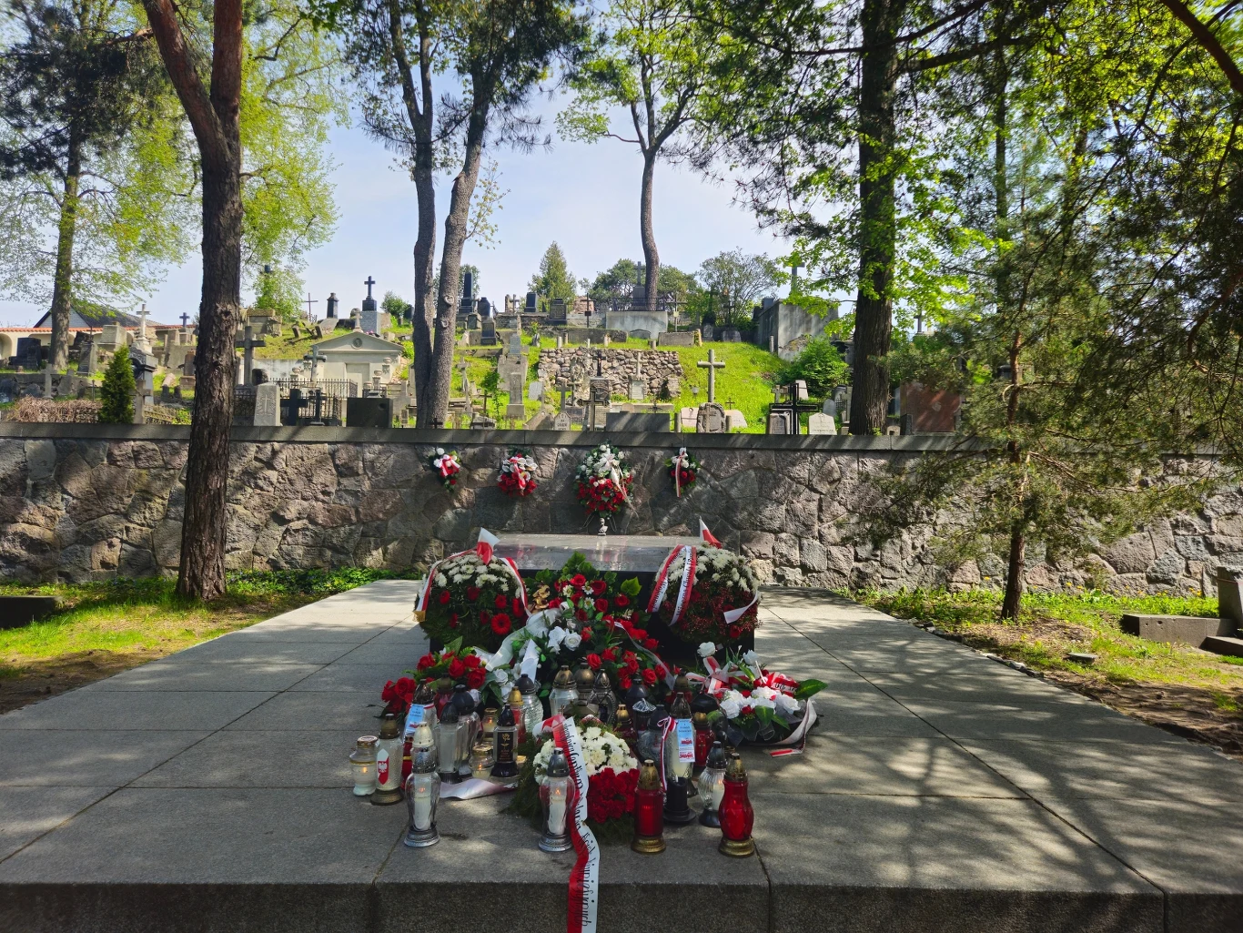 Grób ozdobiony kwiatami i zniczami, otoczony jest wysokimi drzewami. W tle widać mur z krzyżami na cmentarzu, który rozciąga się na wzgórzu. Promienie słońca przenikają przez korony drzew, tworząc spokojną atmosferę.