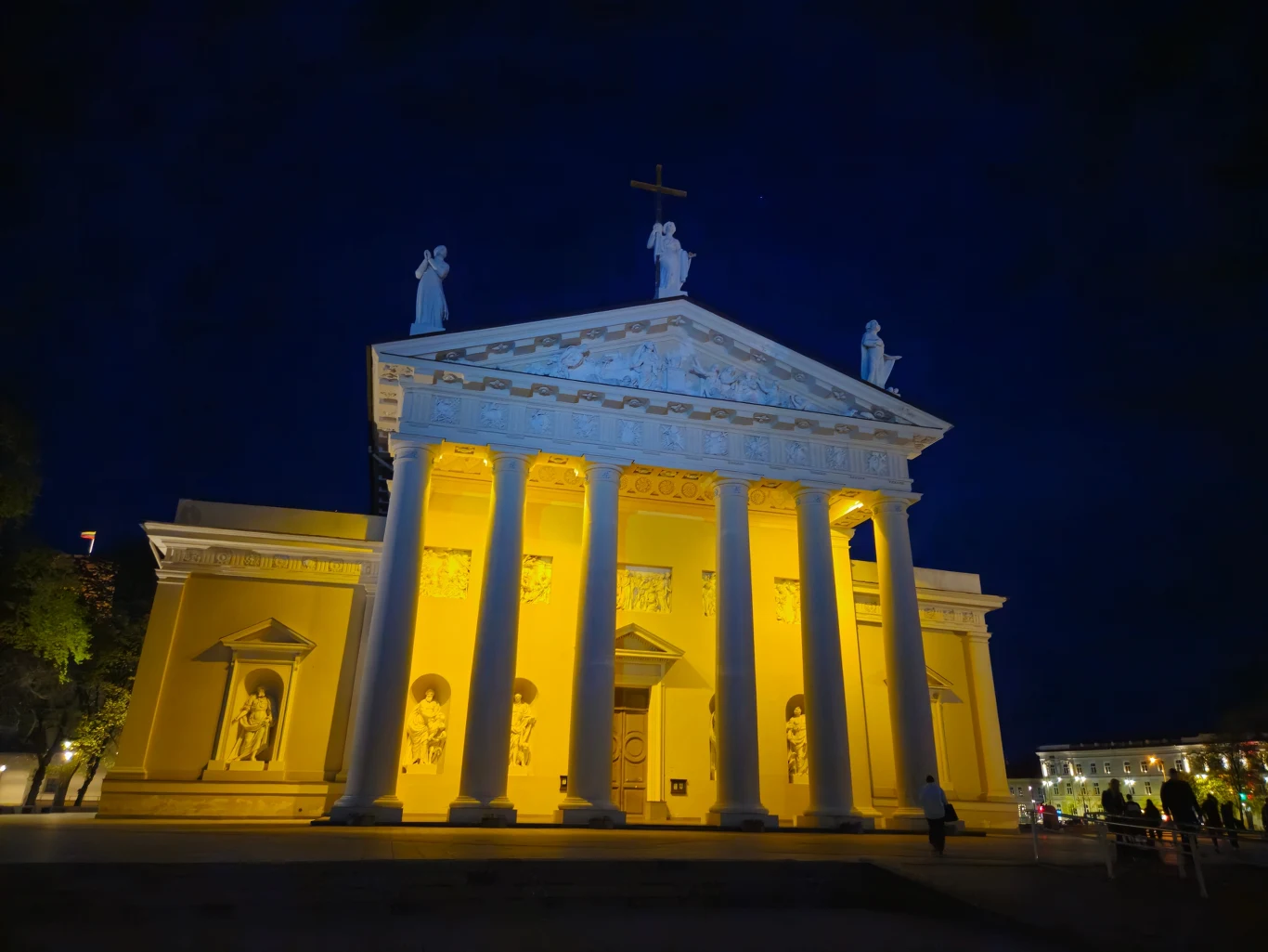 Katedra z imponującą fasadą oświetloną nocą, wyróżniająca się jasnymi kolumnami i rzeźbami na szczycie. Nocne oświetlenie dodaje dramatyzmu monumentalnej strukturze.