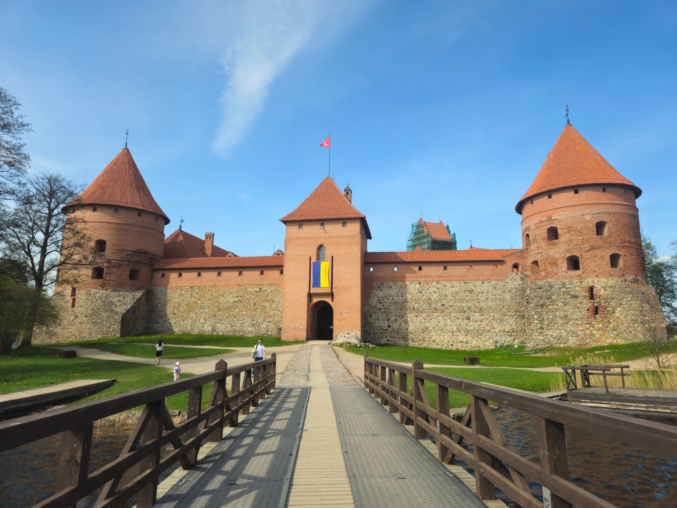 Zamek w Trokach, otoczony fosą, z charakterystycznymi wieżyczkami i czerwonymi dachami, widziany od strony drewnianego mostu.