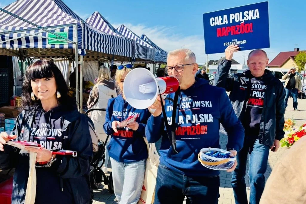 Wybory prezydenckie. Napaść na polityków PO podczas agitacji na rzecz Rafała Trzaskowskiego Grupa osób ubrana w niebieskie bluzy z napisem Trzaskowski cała Polska naprzód! idzie przez targ z budkami w tle. Przewodząca osoba trzyma megafon i koszyk, a inna niesie transparent z tym samym napisem.