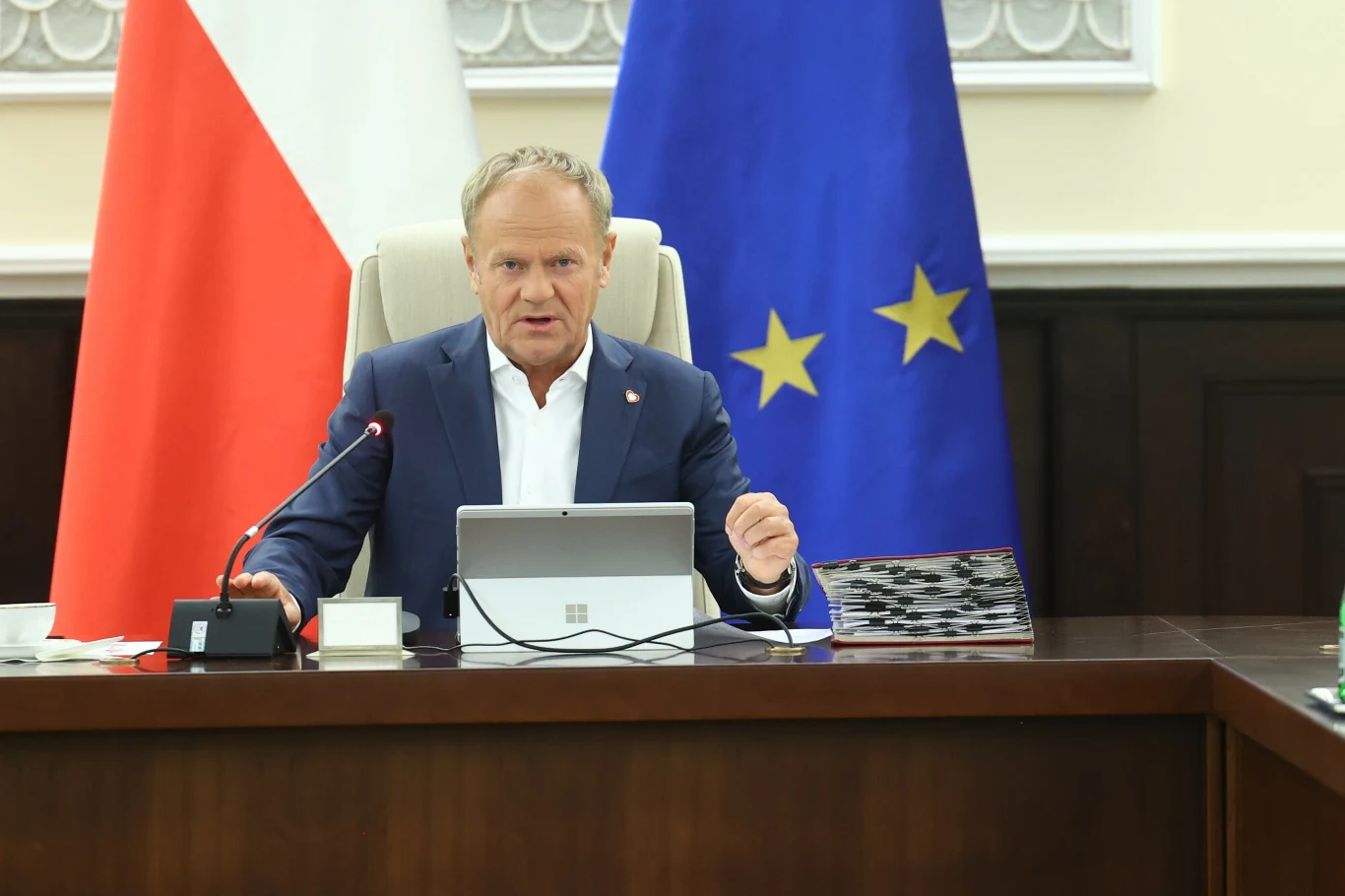 Donald Tusk Mężczyzna siedzi przy stole z flagami Polski i Unii Europejskiej w tle, przed nim znajduje się tablet oraz mikrofon. Mężczyzna jest w średnim wieku, ubrany w marynarkę i koszulę. Wydaje się być w trakcie przemówienia.