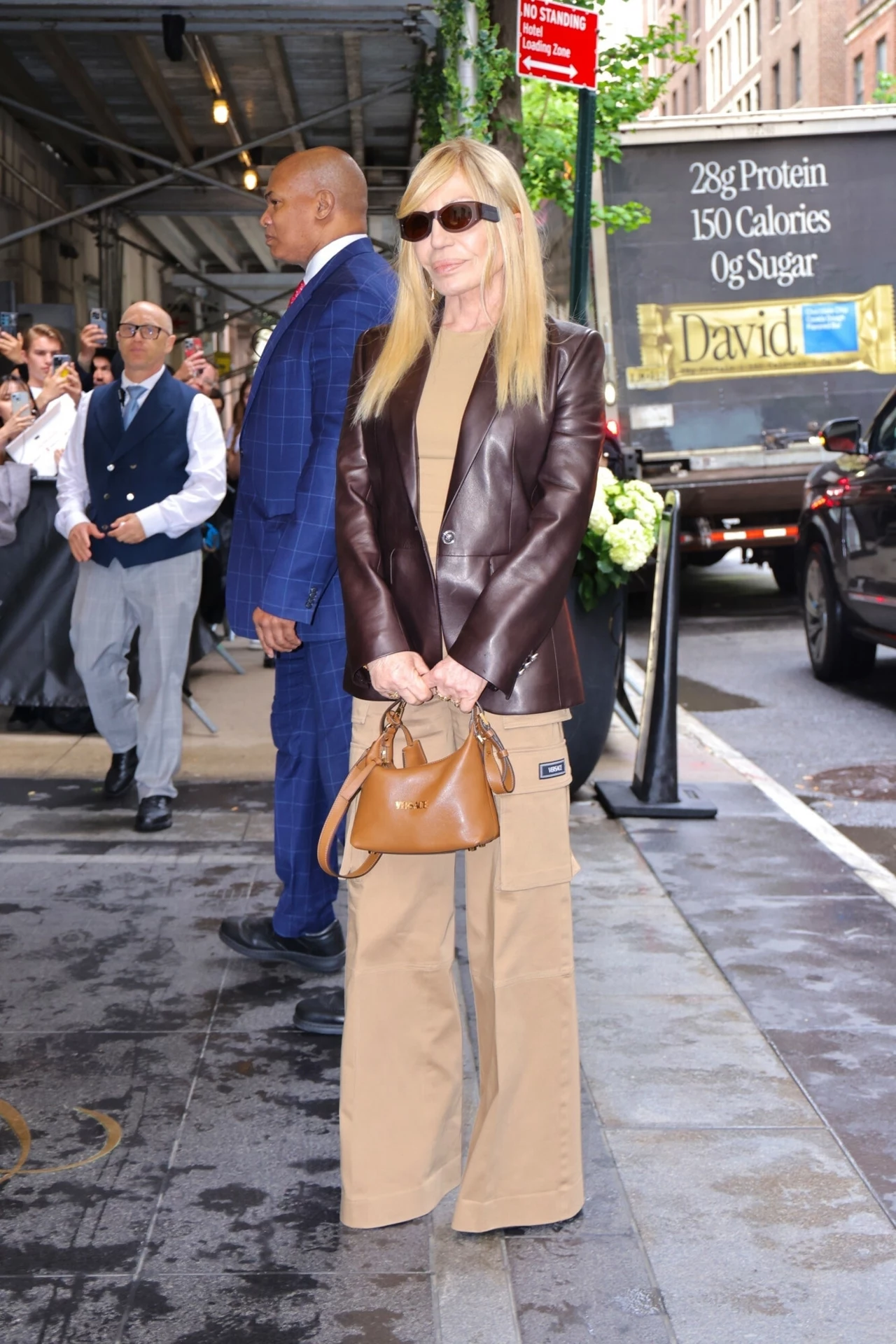 Donatella Versace w luźnej stylizacji Kobieta w okularach przeciwsłonecznych, ubrana w brązową skórzaną kurtkę i beżowe spodnie z torbą, stoi na chodniku w otoczeniu ludzi i samochodów. W tle tablica reklamowa i mężczyzna w niebieskim garniturze.