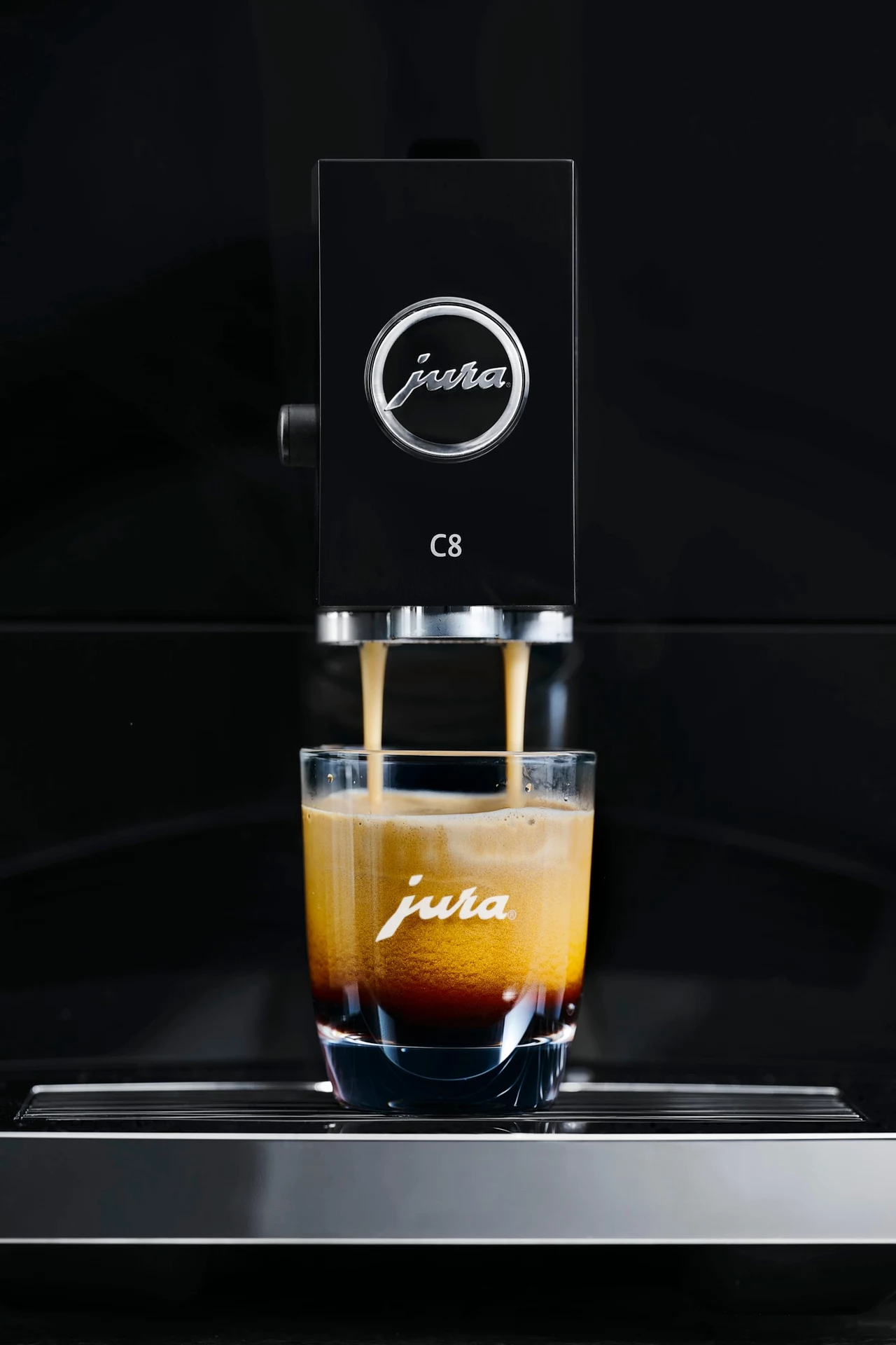 Równomierne zmielenie ziaren jest kluczowe dla jakości naparu Ekspres do kawy Jura C8 przygotowujący porcję espresso. Logo Jura widoczne jest na ekspresie i szklance.