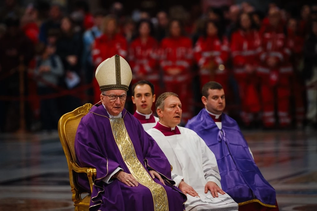 Kardynał Pietro Parolin Mężczyzna w stroju biskupa siedzi na ozdobnym złotym krześle, ubrany w purpurowy ornat z mitrą na głowie. Towarzyszą mu trzej księża w sutannach. W tle widać grupę osób w czerwonych strojach.