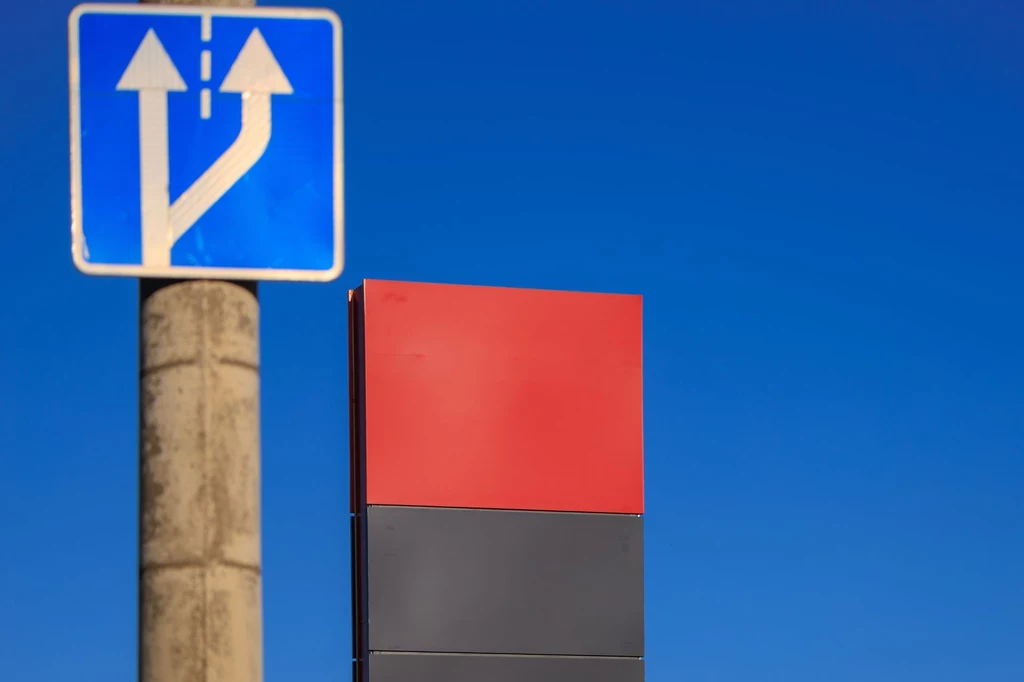 Oszuści zmuszają do zjechania na parking i dogadania się. Znak drogowy na tle czystego, błękitnego nieba. Widać słup z niebieskim znakiem informacyjnym wskazującym rozdzielenie drogi. Obok kolorowa tablica bez widocznych napisów.