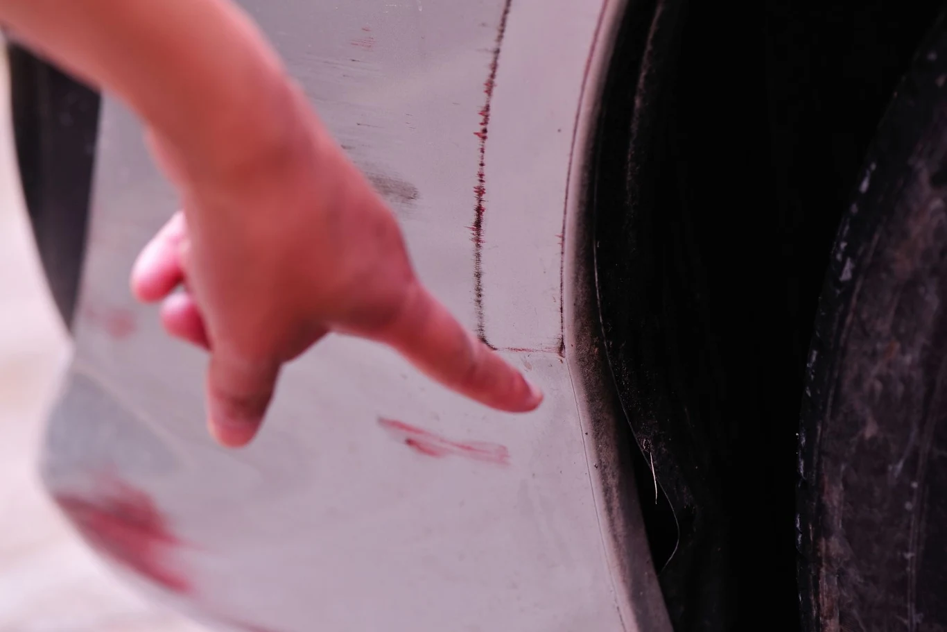 Palec wskazujący dotykający porysowanego zderzaka samochodu z widocznymi zarysowaniami na lakierze.