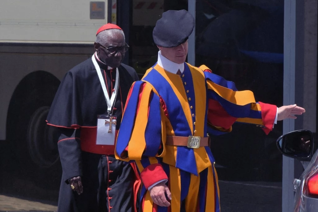 Kardynał Robert Sarah Mężczyzna ubrany w tradycyjny strój Gwardii Szwajcarskiej stoi przed duchownym w czarnej sutannie i czerwonym birecie. W tle widoczny jest pojazd.