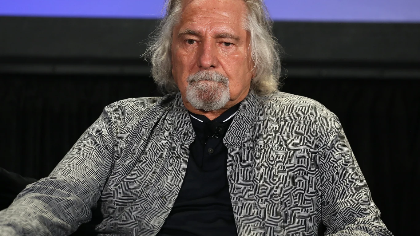 Geezer Butler Starszy mężczyzna o siwych włosach i brodzie siedzi na ciemnym tle. Ma na sobie szarą marynarkę w geometryczny wzór oraz czarną koszulkę. Jego wyraz twarzy jest poważny.