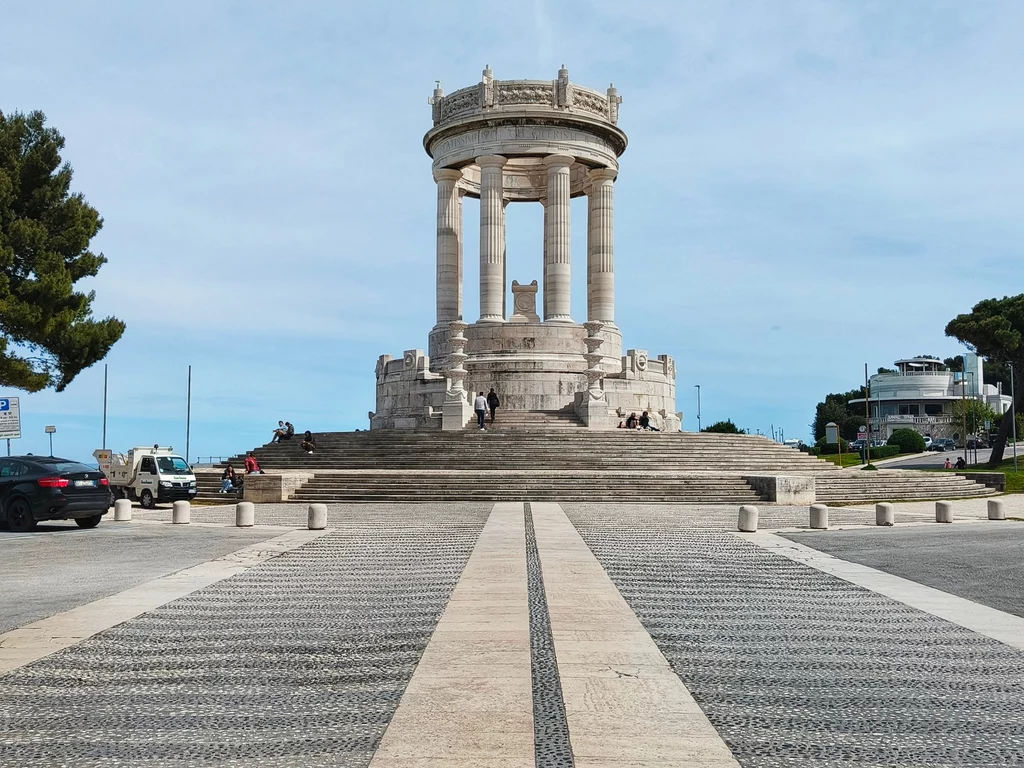 Monumento dei caduti Wysoki starożytny pomnik z kolumnami na szczycie szerokich schodów otoczonych drzewami i budynkami na dalszym planie. Symetryczne ułożenie prowadzące do monumentu, samochody zaparkowane po lewej stronie.