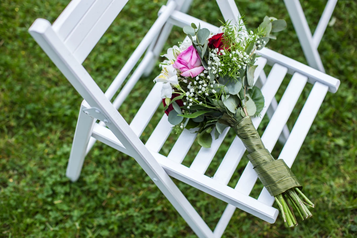 Bukiet kwiatów z różami i zielenią leży na białym składanym krześle ustawionym na trawie.