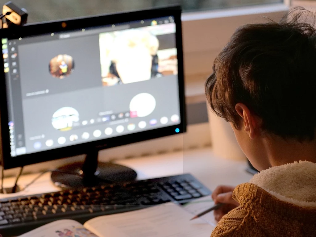 Dziecko siedzi przy biurku, patrząc na monitor komputera, na którym jest włączona wideokonferencja. Obok leży otwarty zeszyt i długopis. Widok zza pleców dziecka podkreśla intymny moment nauki zdalnej czy komunikacji.
